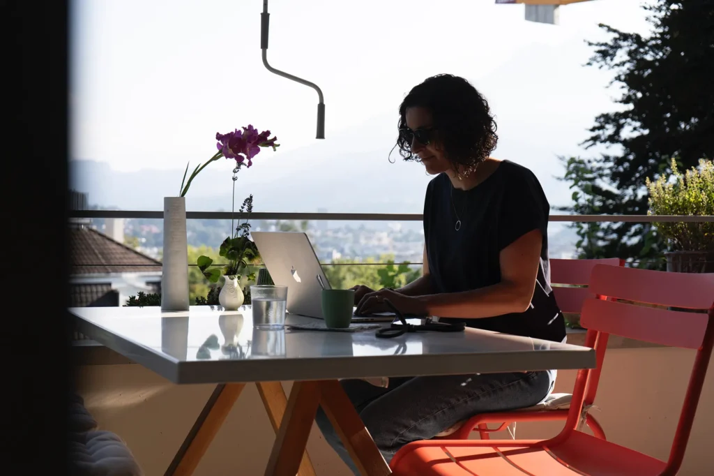 frau-balkon-terrasse-aussicht-laptop-blume-vase-wasserglas-rote-gartenstuehle