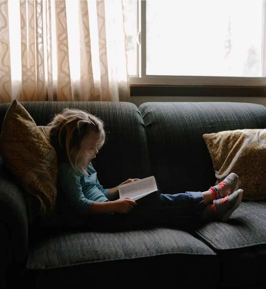 kind-buch-sofa-lesen-kissen-vorhang-fenster